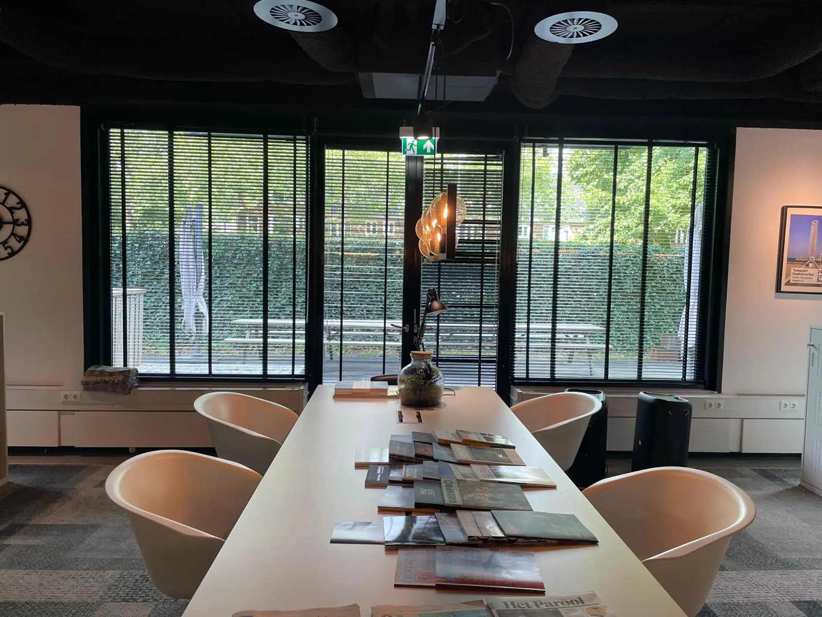 Moderne vergaderruimte aan de De Cuserstraat met een lange tafel, designstoelen en tijdschriften, met uitzicht op een groene tuin door grote ramen met jaloezieën.